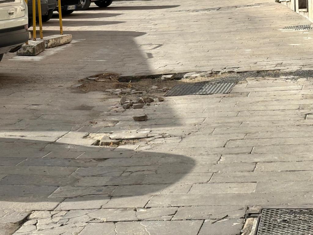 Uneven cobblestone street with a hole and scattered debris, surrounded by parked cars.
