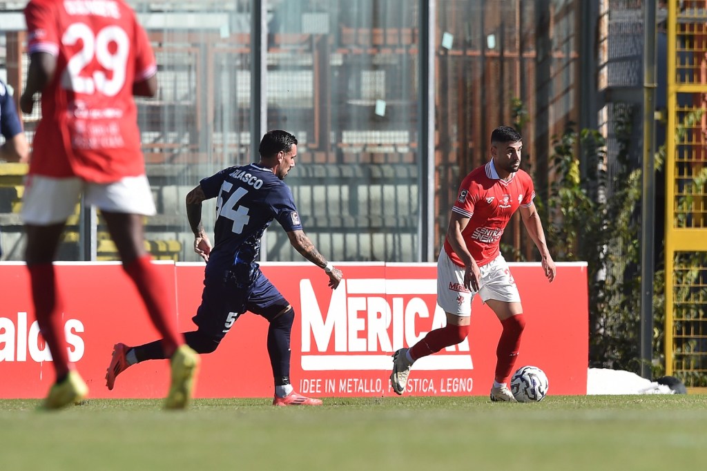 Giocatori di calcio in azione durante una partita tra Perugia e Rimini, con un calciatore in maglia rossa che dribbla un avversario in maglia blu.