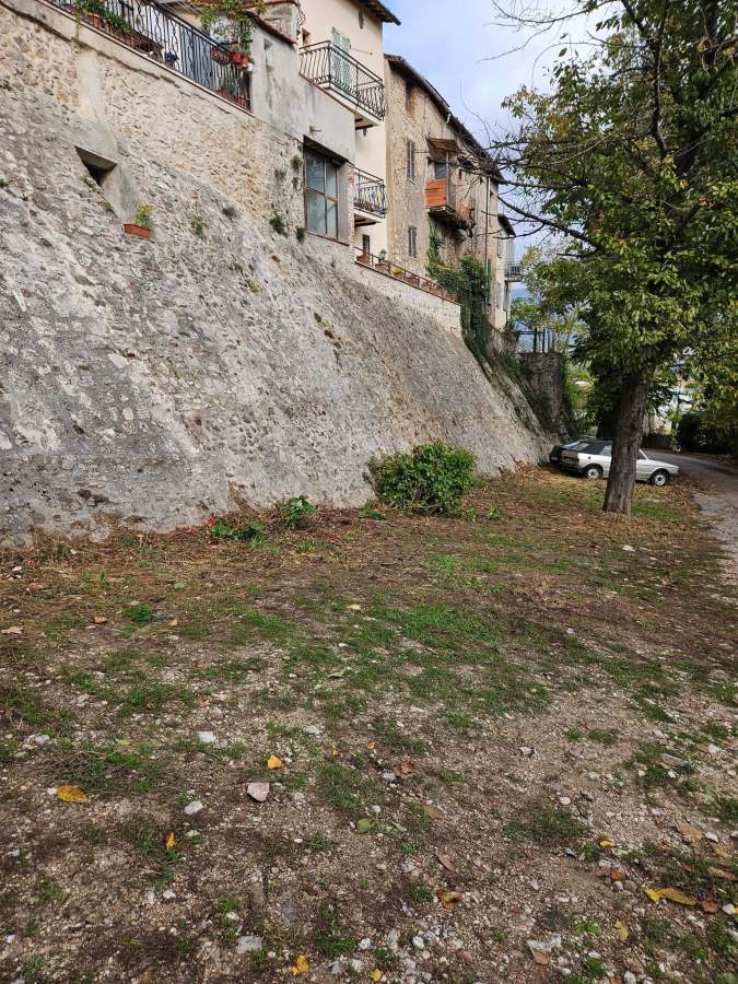 Vista delle antiche mura di Collescipoli, ripulite da erbacce e rifiuti, con case storiche sullo sfondo e un'area verde recentemente curata.