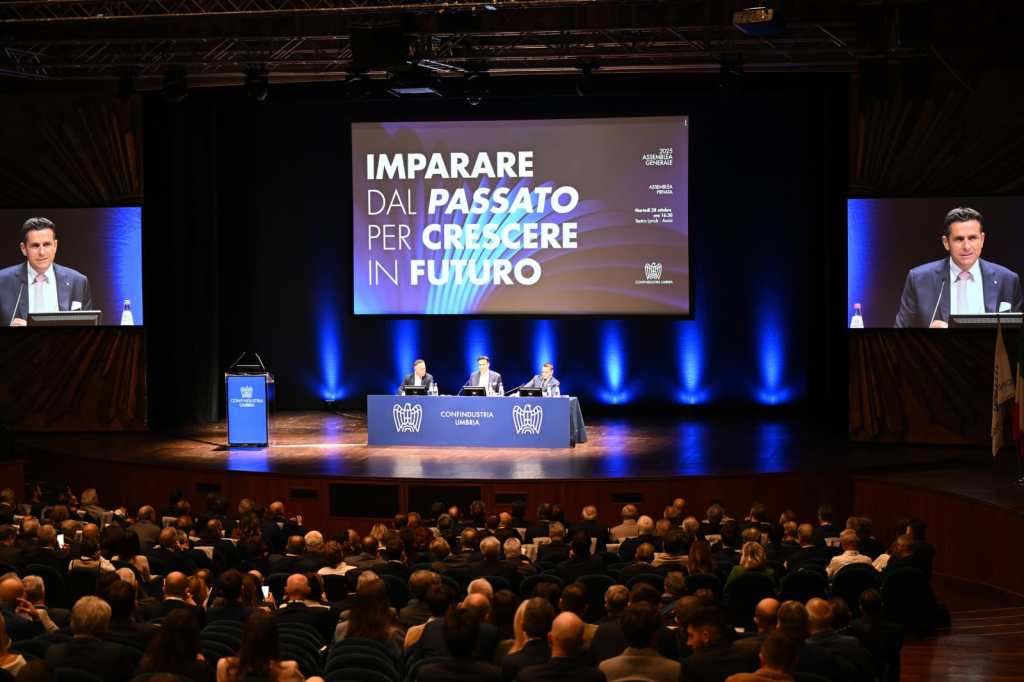 Vista dell'Assemblea Generale di Confindustria Umbria al Teatro Lyrick di Assisi, con relatori al tavolo e un pubblico in sala. Sullo sfondo, uno schermo mostra il tema 'Imparare dal passato per crescere in futuro'.