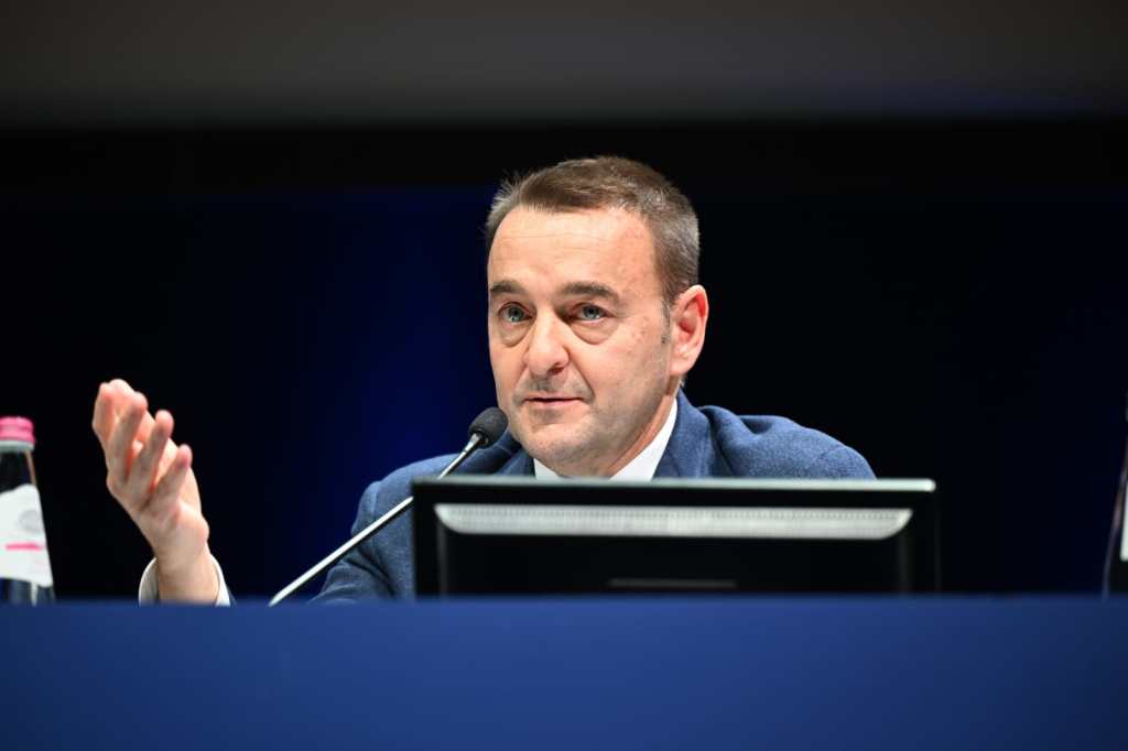 Un uomo parla al microfono durante un'assemblea, con un computer portatile e una bottiglia d'acqua sul tavolo.