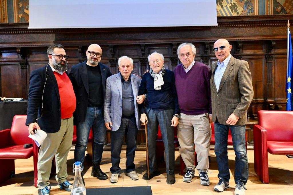 A group of six men standing together on stage at a conference, some wearing glasses, with a mix of casual and formal attire, in front of a wooden backdrop.