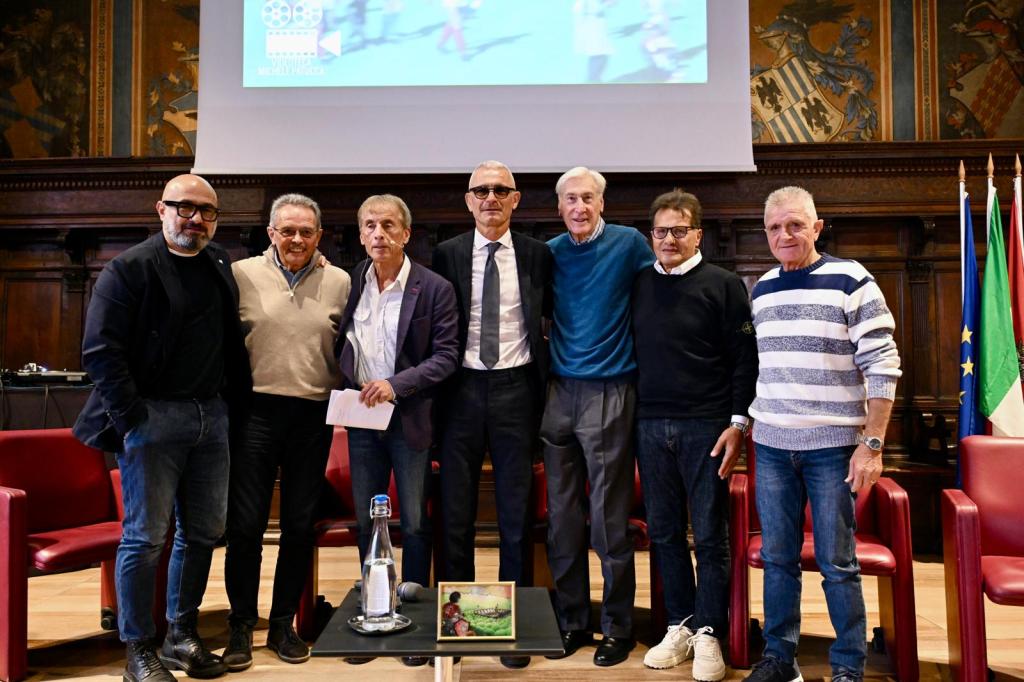 A group of seven men standing together in a formal setting, with several wearing glasses and others in casual attire, in front of a screen displaying a colorful image.