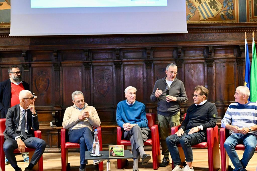 A panel of six men seated in red chairs during a discussion event, with a wooden backdrop and flags in the background.