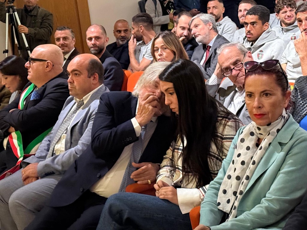 A group of attendees seated at an event, featuring various individuals in formal attire, including a woman with long hair and another woman in a light blue coat with a polka dot scarf, while expressions of curiosity and engagement are visible.