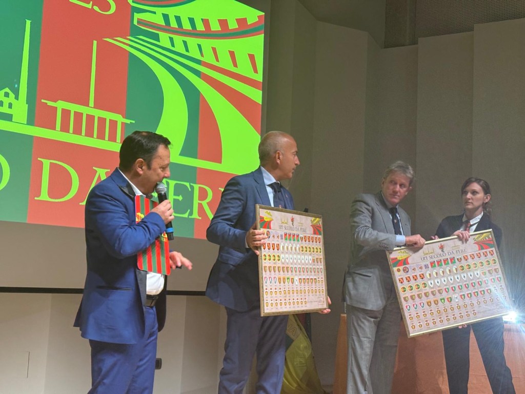 Cerimonia di apertura dei festeggiamenti per il centenario della Ternana Calcio, con relatori che mostrano tabelle commemorative su un palco, dietro di loro è visibile uno sfondo colorato con il logo della Ternana.