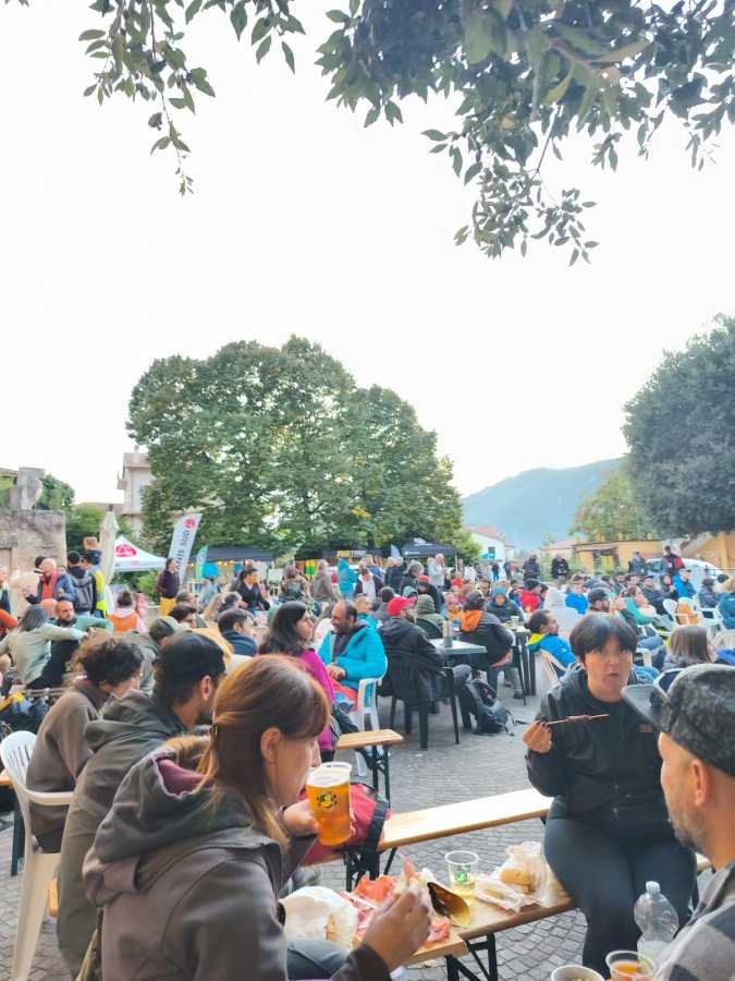 Un gruppo di persone riunite in un piazza durante un festival, con tavoli, sedie, e cibo. Alcuni partecipanti stanno mangiando e bevendo, mentre altri sono coinvolti nella conversazione. Sullo sfondo, si vedono tende e alberi.