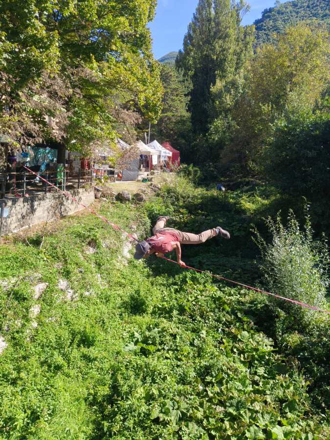 Un partecipante fa slackline sopra un letto di vegetazione in una competizione outdoor a Ferentillo, con tende e alberi sullo sfondo.