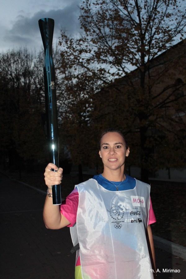 A woman holding a torch in a park during twilight, wearing a white vest with Olympic-themed logos.