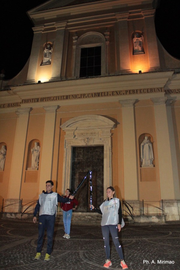 Two people in sports attire holding illuminated objects in front of a lit building at night, with a third person in the background.