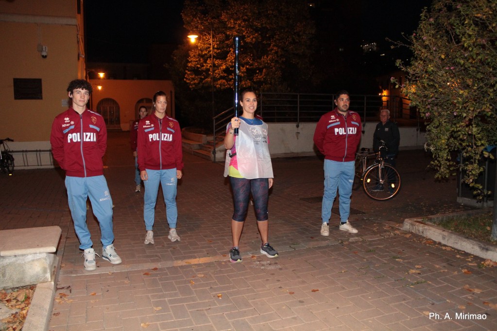 A group of four individuals in athletic wear, two men and two women, are gathered in an outdoor setting at night. One woman is holding a lighted baton, while the others stand in a supportive stance, wearing uniforms labeled 'Polizia.' Trees and urban lighting are visible in the background.