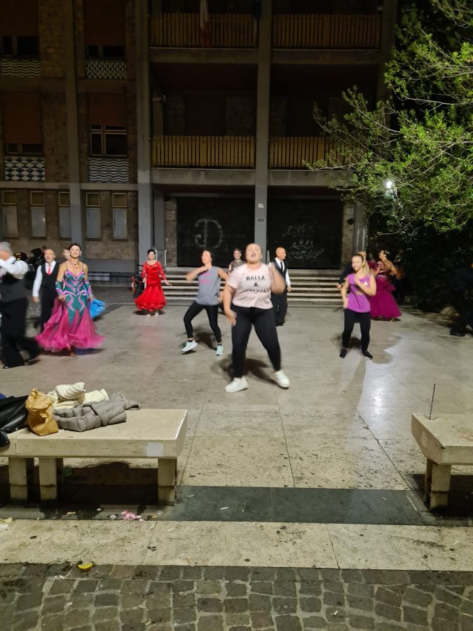 Gruppo di ballerini in abiti colorati si esibiscono in una piazza, con panchine e vegetazione circostante.