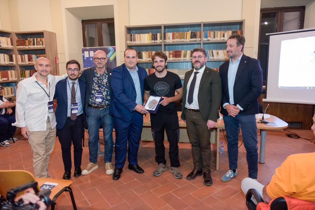Un gruppo di sei persone sorridenti posano insieme all'interno di una sala biblioteca, con scaffali di libri sullo sfondo, mentre uno di loro tiene un premio o un riconoscimento.