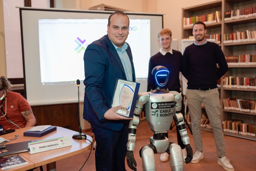 A group of people, including a man holding an award and a humanoid robot, pose for a photo in a library setting during an event focused on technology and society.