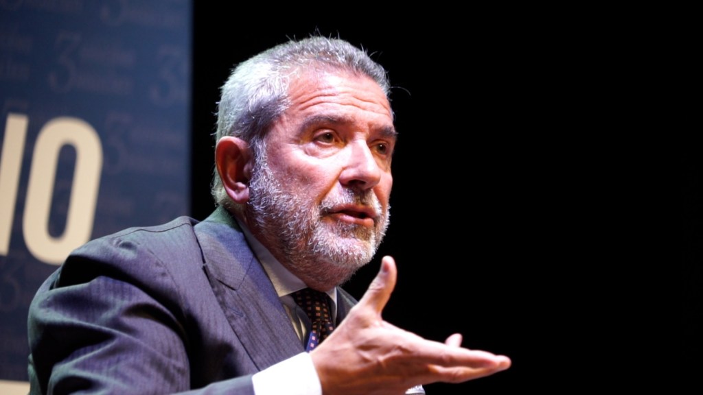 Un oratore maschio con barba grigia e capelli bianchi, vestito in un abito scuro, parla durante un evento politico, con un'espressione seria e gesti delle mani visibili sul palco.