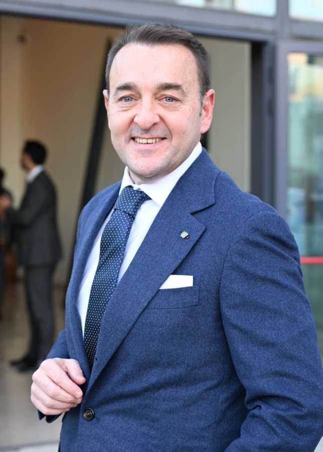 Giammarco Urbani sorridente, indossando un abito blu, mentre si trova davanti a una porta in un contesto professionale.