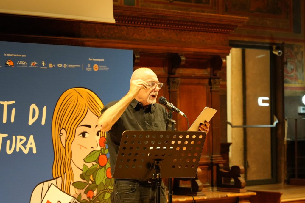 Un uomo legge un testo al microfono durante l'evento UmbriaLibri 2025, davanti a un pubblico in una sala affollata, con un grande pannello illustrato sullo sfondo.