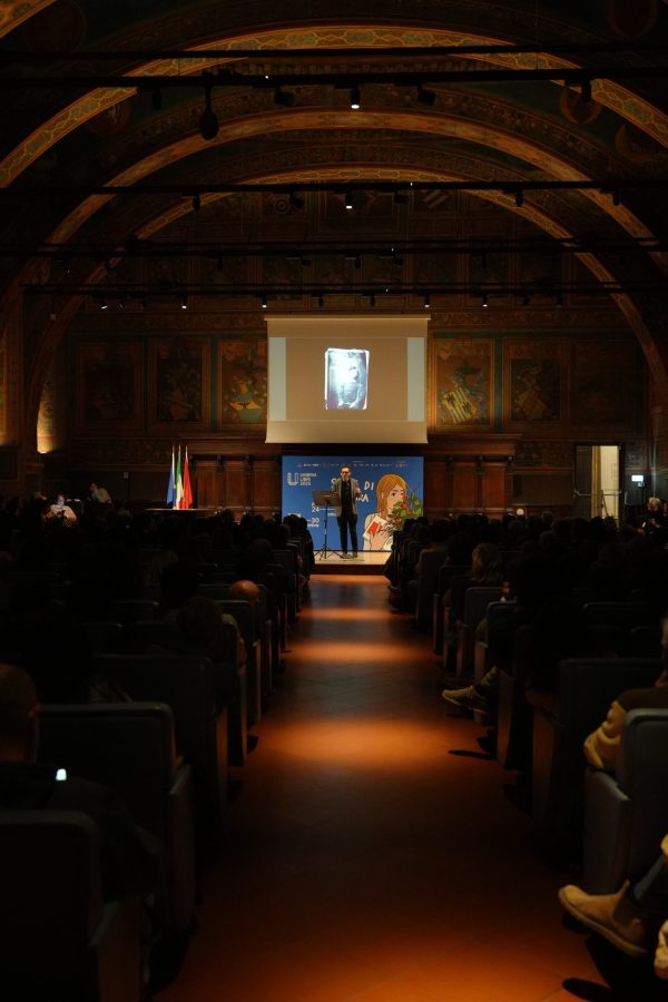 Un relatore parla su un palco illuminato, davanti a un pubblico in un'ampia sala. Sullo sfondo, un'immagine proiettata e bandiere italiane si vedono ai lati. La scena è allestita per un evento culturale durante il festival UmbriaLibri 2025.