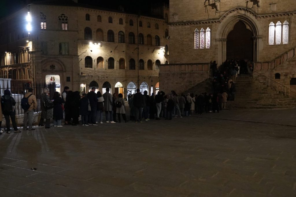 Una lunga fila di persone che si snodano davanti a un edificio storico illuminato, probabilmente in attesa di un evento culturale o letterario.