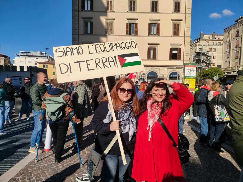 Due donne sorridenti partecipano a una manifestazione con un cartello che dice 'SIAMO L'EQUIPAGGIO DI TERRA', sullo sfondo si vedono altre persone e edifici.