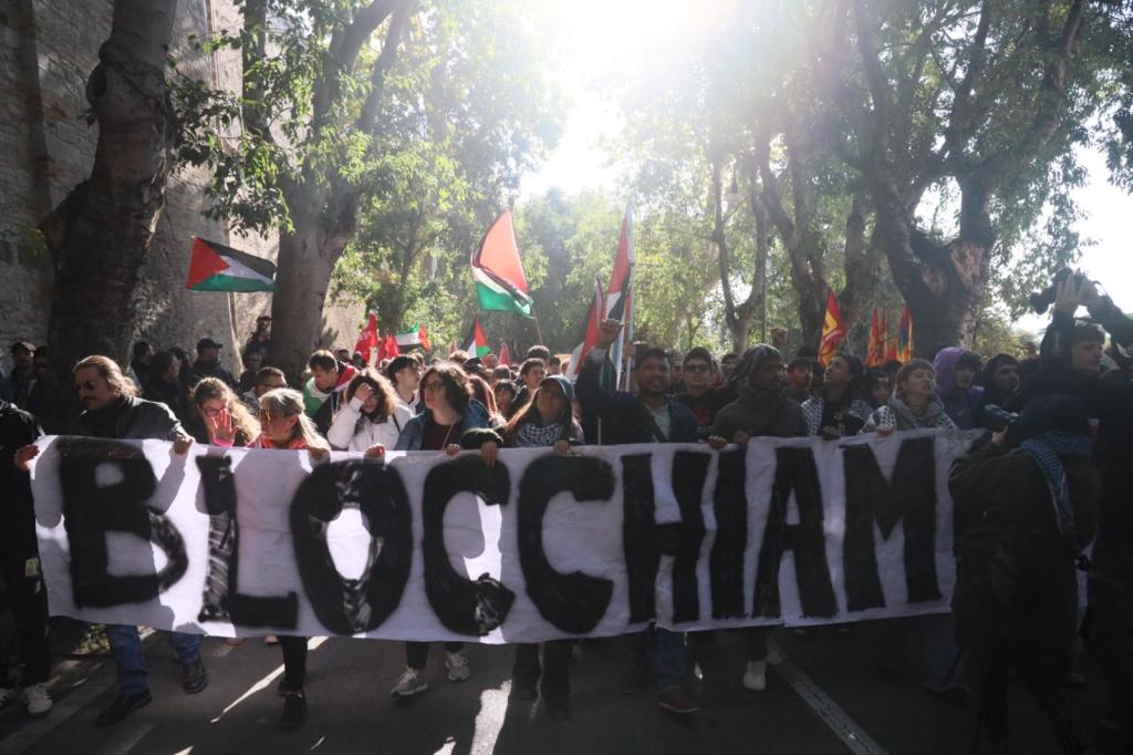 Manifestazione di protesta a Perugia con manifestanti che sventolano bandiere palestinesi e portano uno striscione con la scritta 'BLOCCIAMO'.
