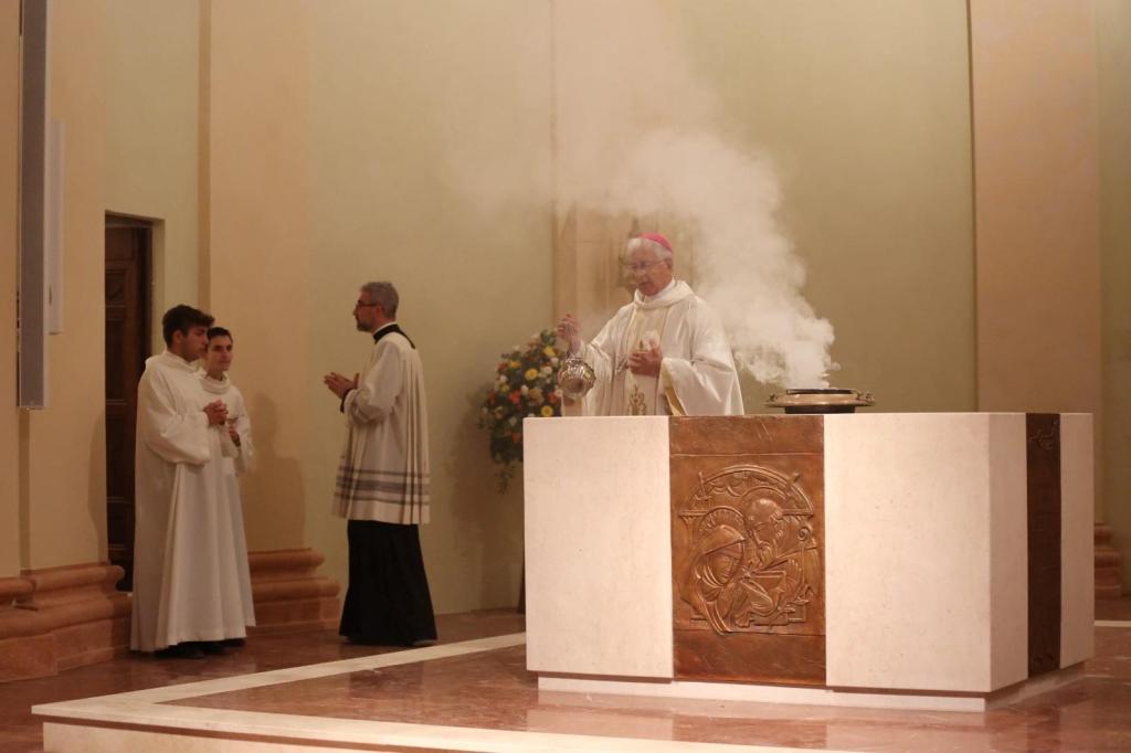 Un arcivescovo in abito liturgico benedice l'altare durante una cerimonia religiosa, mentre due giovani assistenti e un sacerdote sono presenti accanto a lui. Una colonna di fumo di incenso si alza dall'altare.