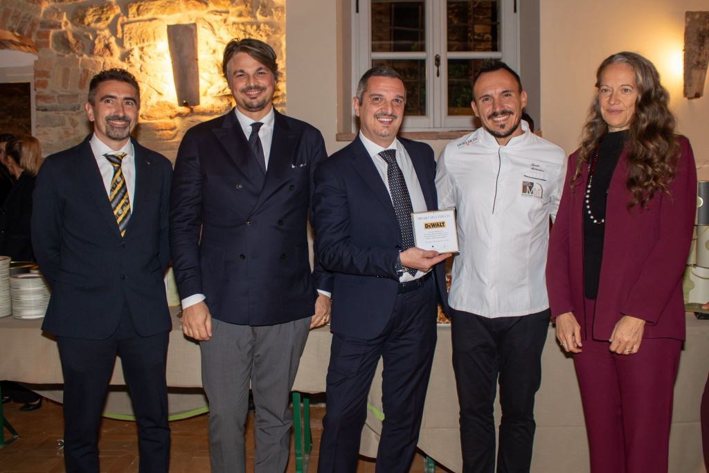 Quattro uomini e una donna in abbigliamento elegante durante un evento di gala, con un premiato che tiene un trofeo, sullo sfondo decorativo di un ristorante.