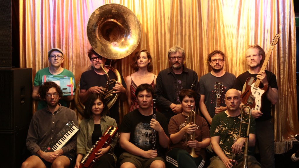 Un gruppo di musicisti posa di fronte a una tenda dorata, con vari strumenti musicali come chitarre, trombe e una tuba.