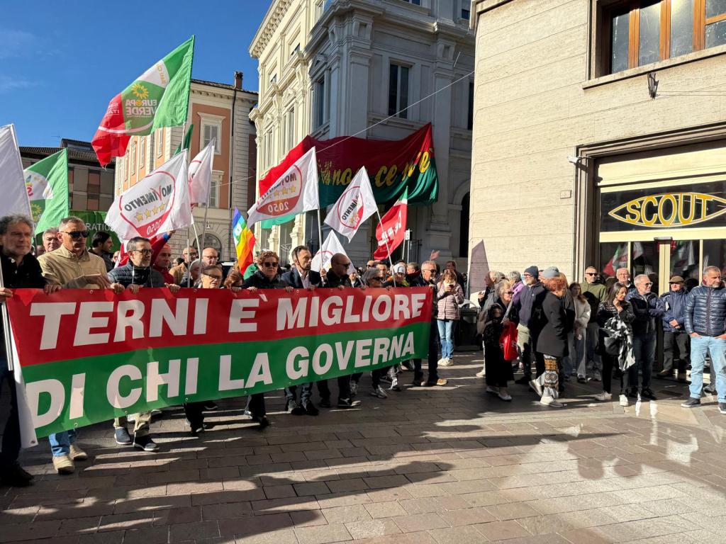 Un gruppo di manifestanti in piazza, con uno striscione che recita 'TERNI È MIGLIORE DI CHI LA GOVERNA', accompagnato da bandiere e un numeroso pubblico in un ambiente cittadino.