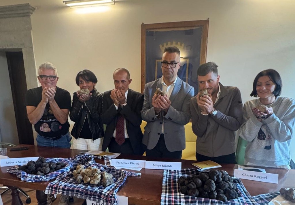 Un gruppo di sei persone in una conferenza stampa, tutte con tartufi nelle mani e sorridenti, con tavolo di tartufi in primo piano.