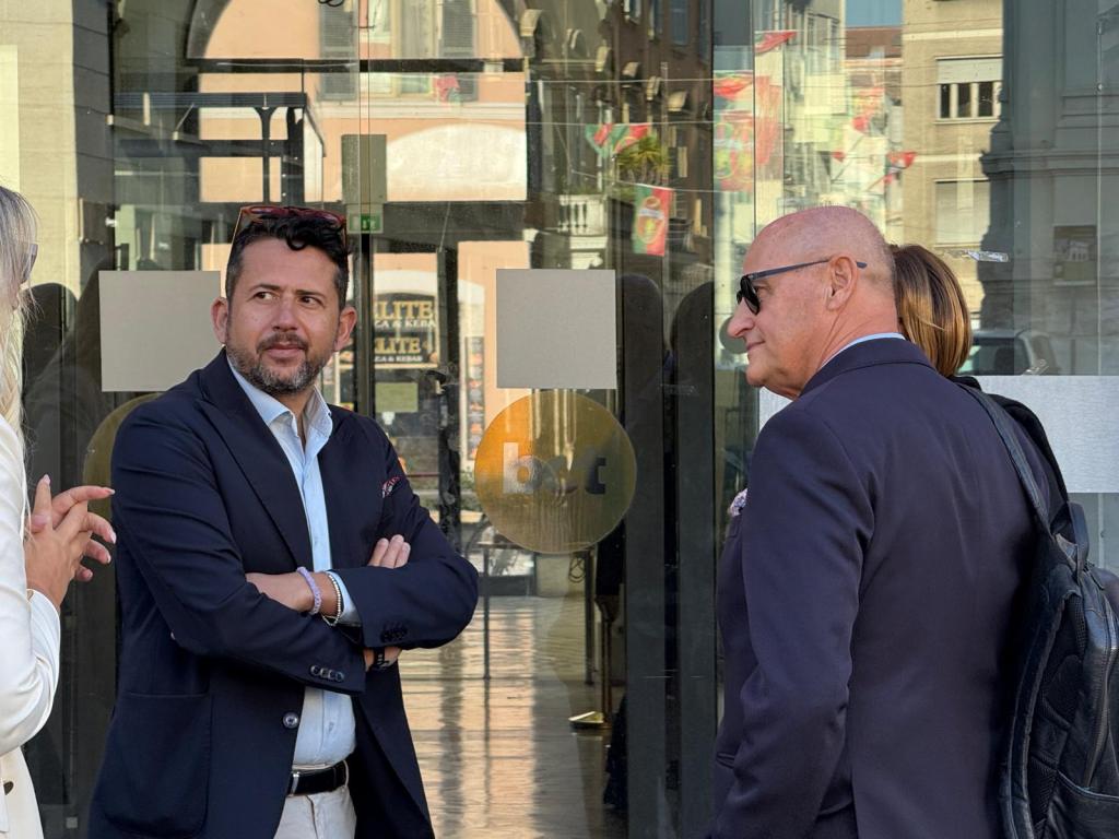 A group of professionals standing outside a building with glass doors, engaging in conversation. One man wearing a blazer is listening attentively, while another man, also in a suit, is visible from the side. A woman in a light-colored outfit is gesturing as she speaks.