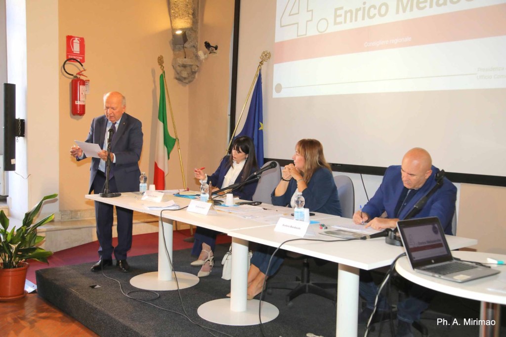 Un incontro ufficiale con un relatore al microfono e membri del pubblico seduti a un tavolo. Sullo sfondo si vedono bandiere italiane ed europee.