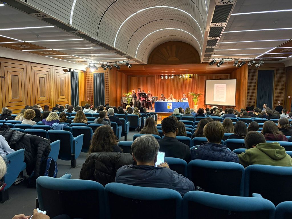 Un evento pubblico in una sala con un ampio pubblico seduto, mentre un gruppo di relatori è in primo piano su un palco con un tavolo e una presentazione visiva.