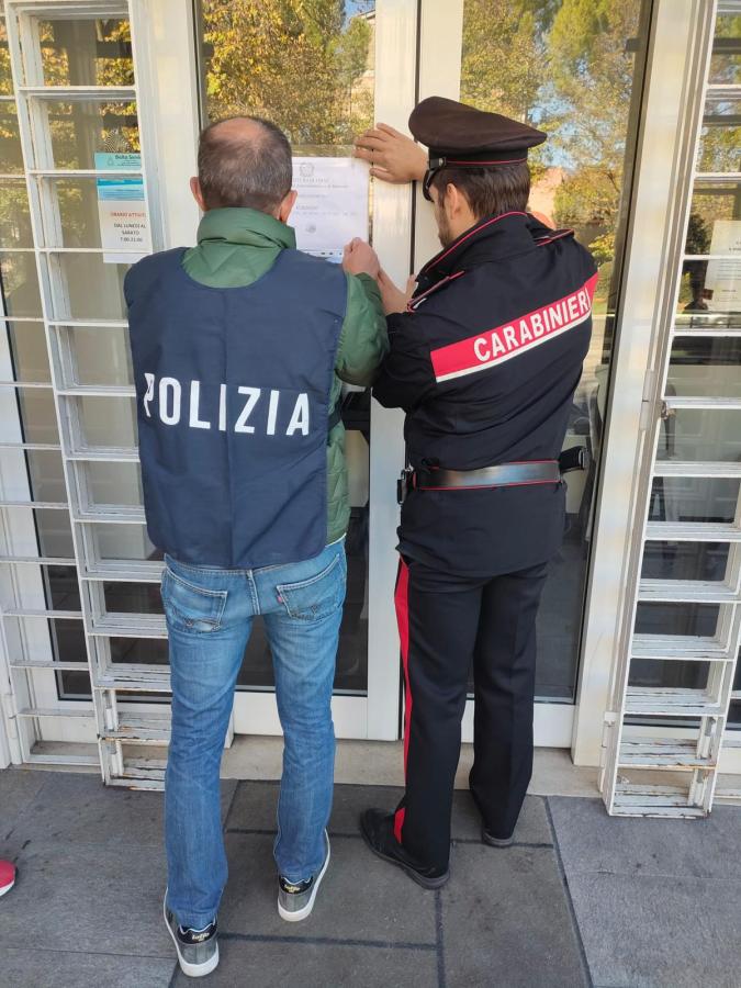 Two law enforcement officers are posting a notice on a door. One officer is wearing a 'Polizia' vest and the other is in a 'Carabinieri' uniform.