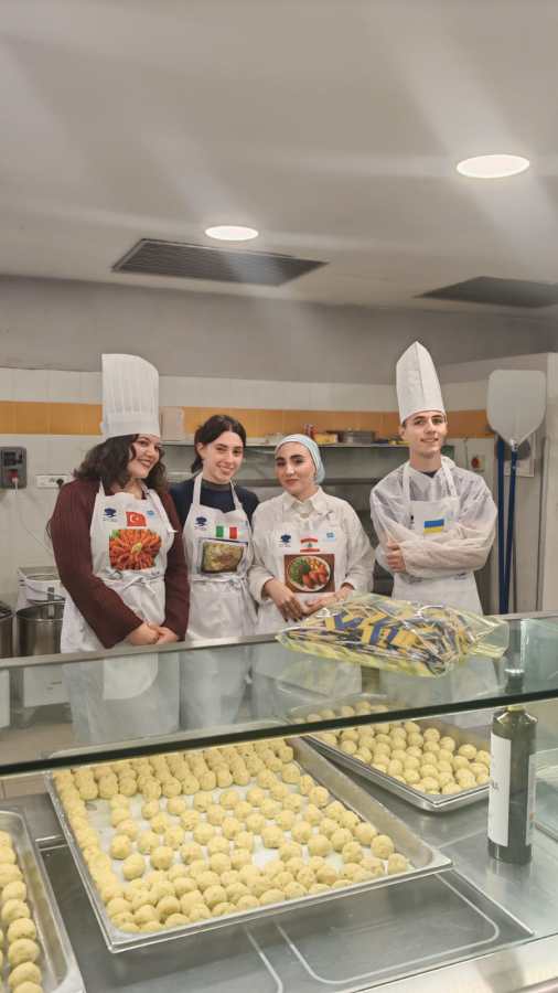 Quattro studenti in uniforme da cucina sorridono insieme in una cucina, con piatti tradizionali davanti a loro, rappresentando diverse culture gastronomiche.