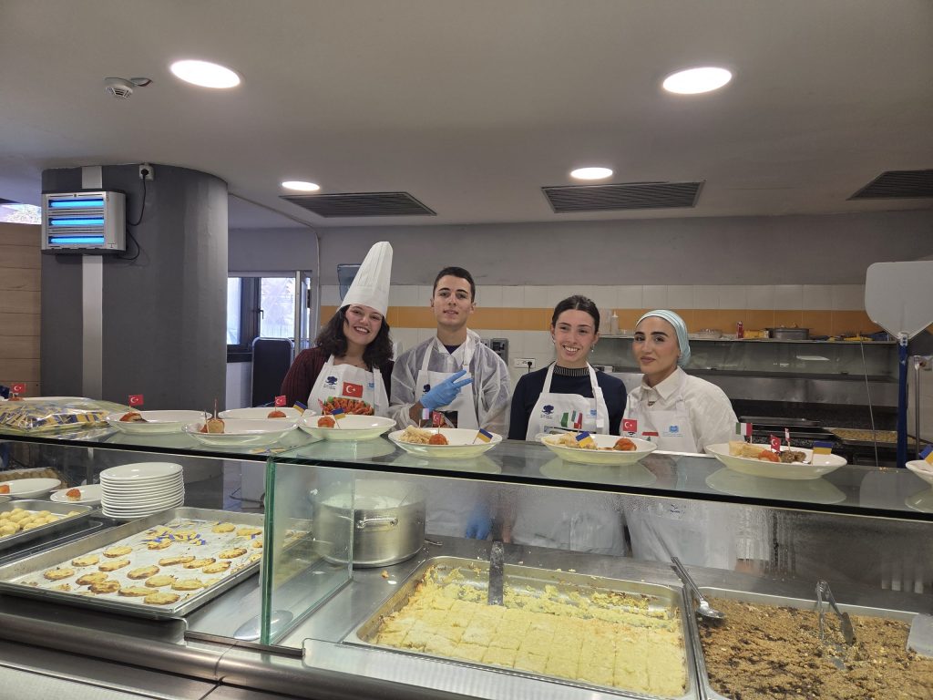 Quattro studenti sorridenti vestiti con divise da chef davanti a un bancone di cucina, con piatti e decorazioni tipiche di diverse culture gastronomiche.