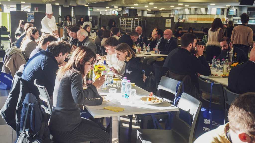 Un gruppo di persone sedute a un tavolo durante il concorso gastronomico "Da leccarsi i baffi", mentre degustano diversi piatti e interagiscono tra loro.