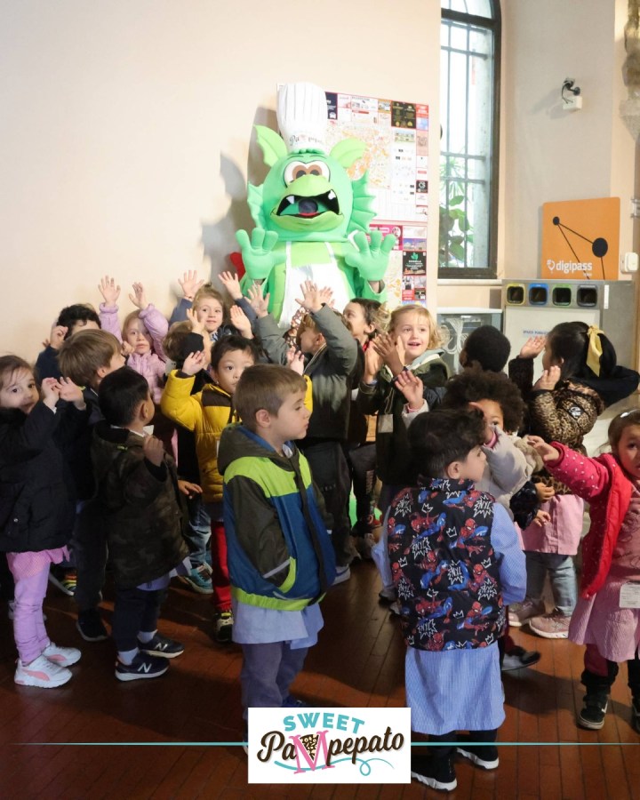 Un gruppo di bambini sorridenti che interagiscono con una mascotte verde a forma di mostro chef, durante l'evento Sweet Pampepato a Terni.