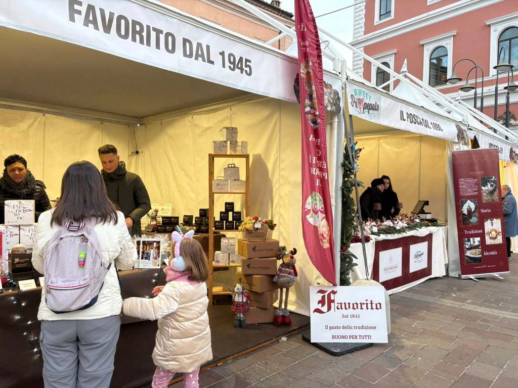 A busy market scene featuring a booth labeled 'Favorito dal 1945' with people interacting and browsing products. A woman and a child are in front of the booth, while another vendor is behind the counter.