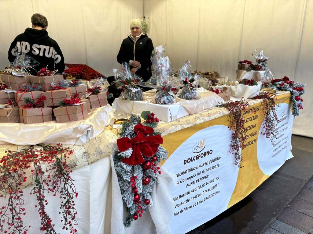 A market stall decorated for the holidays, displaying various gift boxes and festive arrangements. Two individuals are present, one standing behind the counter and another in the background. The stall features a sign for 'DOLCEFORNO' with contact information.