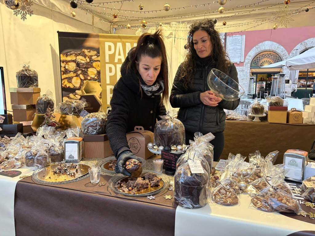 Two women at a chocolate stall during a market event. One woman is arranging chocolate products while the other holds a bowl. The display includes various packaged chocolates and pastries, set against a festive backdrop.