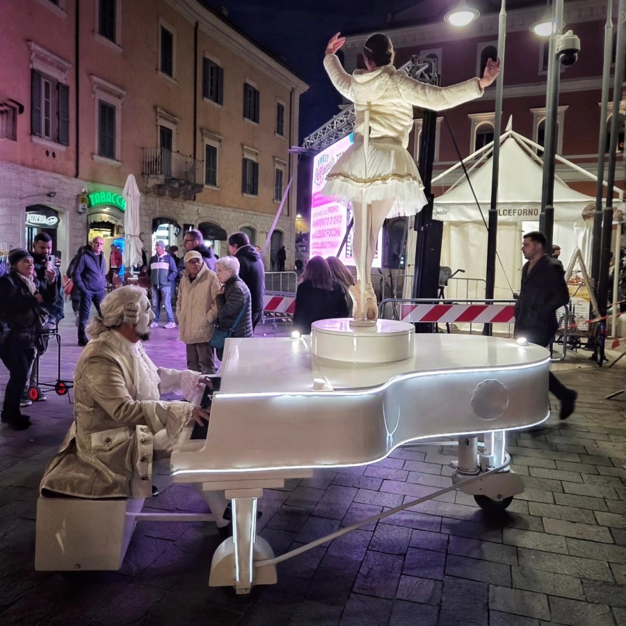 A man in a historical costume playing a white illuminated piano, while a ballerina in a white outfit performs on top, with an audience watching in a nighttime city square.