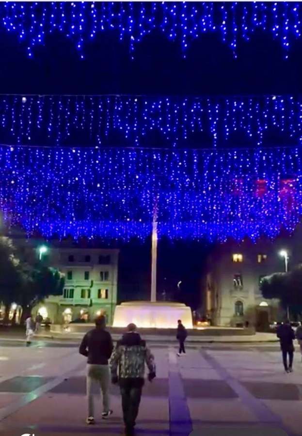A night view of a square adorned with blue decorative lights overhead, with people walking towards a fountain in the center.