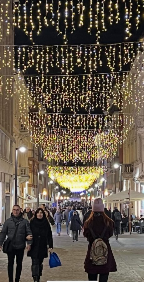 A night scene of a bustling street adorned with hanging lights of various colors, with people walking down the center and cafes visible on the sides.