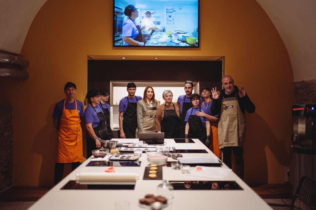 Gruppo di giovani lavoratori de 'Il gusto che unisce' in un laboratorio di pasticceria, sorridendo e indossando grembiuli, con utensili da cucina e un grande schermo alle spalle.