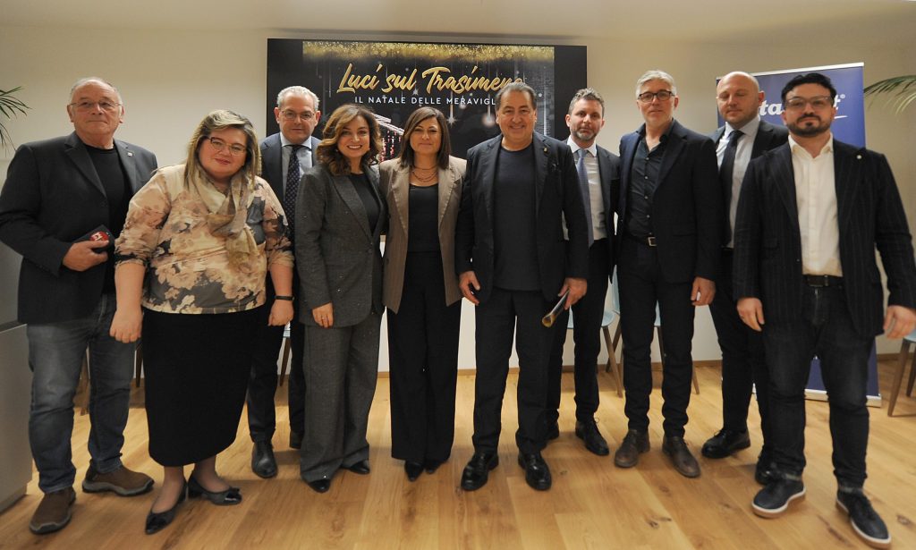 Gruppo di persone in posa durante la presentazione dell'evento 'Luci sul Trasimeno', con sfondo decorato e stendardi.