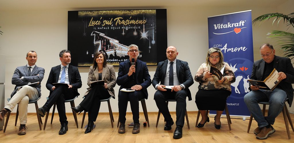 Un gruppo di relatori partecipa a una conferenza stampa per presentare l'evento 'Luci sul Trasimeno', con un grande cartello sullo sfondo che evidenzia il tema natalizio.