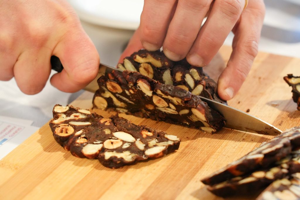Una persona che affetta un dolce tradizionale chiamato Pampepato su un tagliere di legno, mostrando pezzi di nocciole e mandorle inclusi nel dessert.