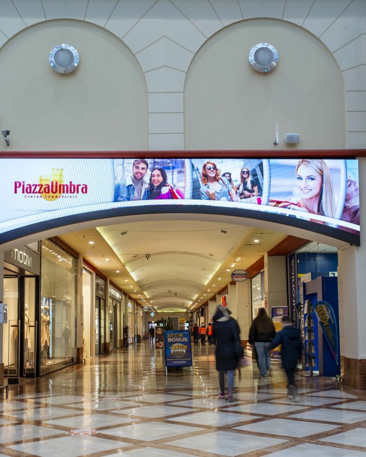 Interno del centro commerciale PiazzaUmbra a Trevi, con un grande schermo pubblicitario sopra l'ingresso che mostra immagini di persone sorridenti. Diversi negozi sono visibili lungo il corridoio.