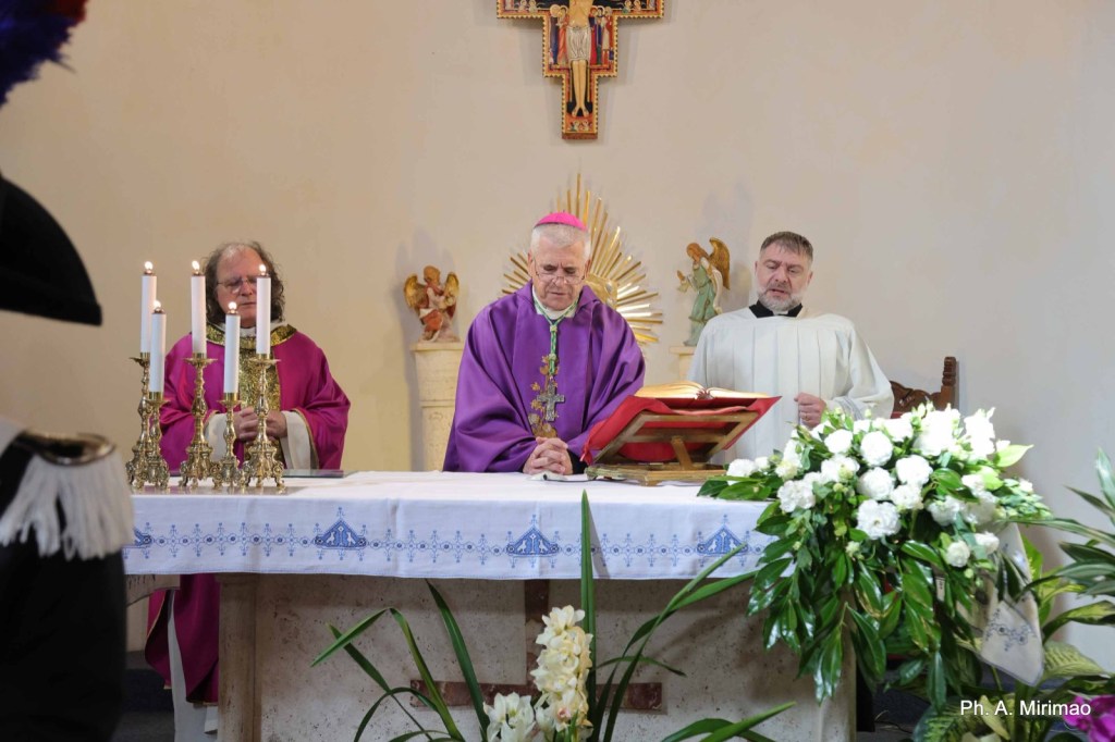 A religious ceremony taking place at an altar, with a bishop and two clergy members dressed in liturgical vestments, surrounded by candles and floral arrangements.